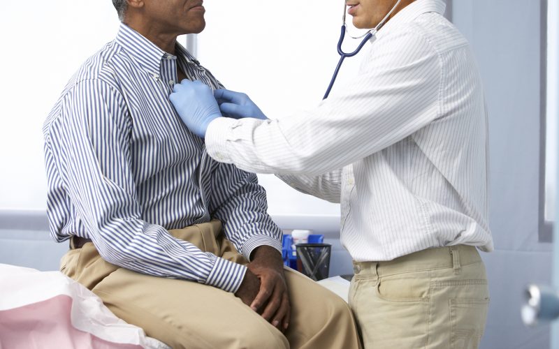 Doctor In Surgery Listening To Male Patient's Chest Using A Stethoscope.